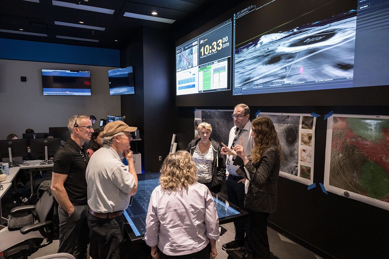 A group of people are in a room with large screens on the walls that display lunar mission information. There are lunar maps on the walls under the screen. The group of seven people stands around an electronic touch screen table that displays a map of the lunar surface. The group is listening to a woman on the right side of the table as she describes something.